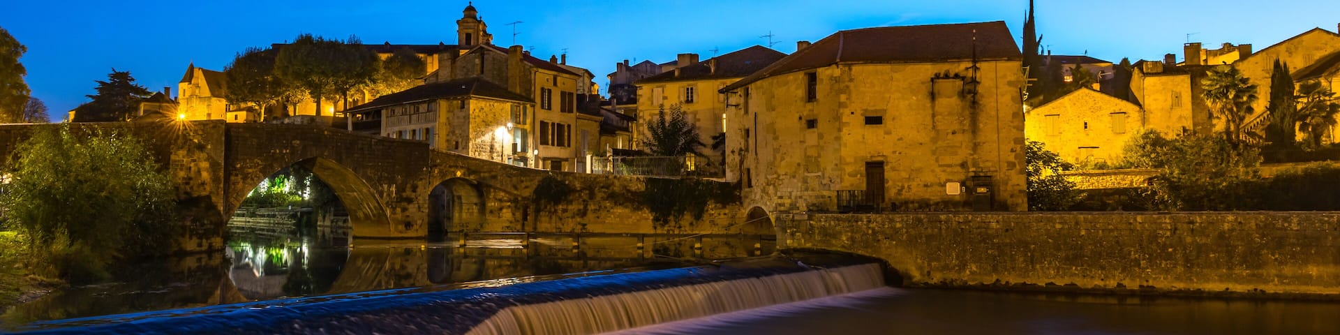 A river with a waterfall, stone bridge, and illuminated buildings at dusk.