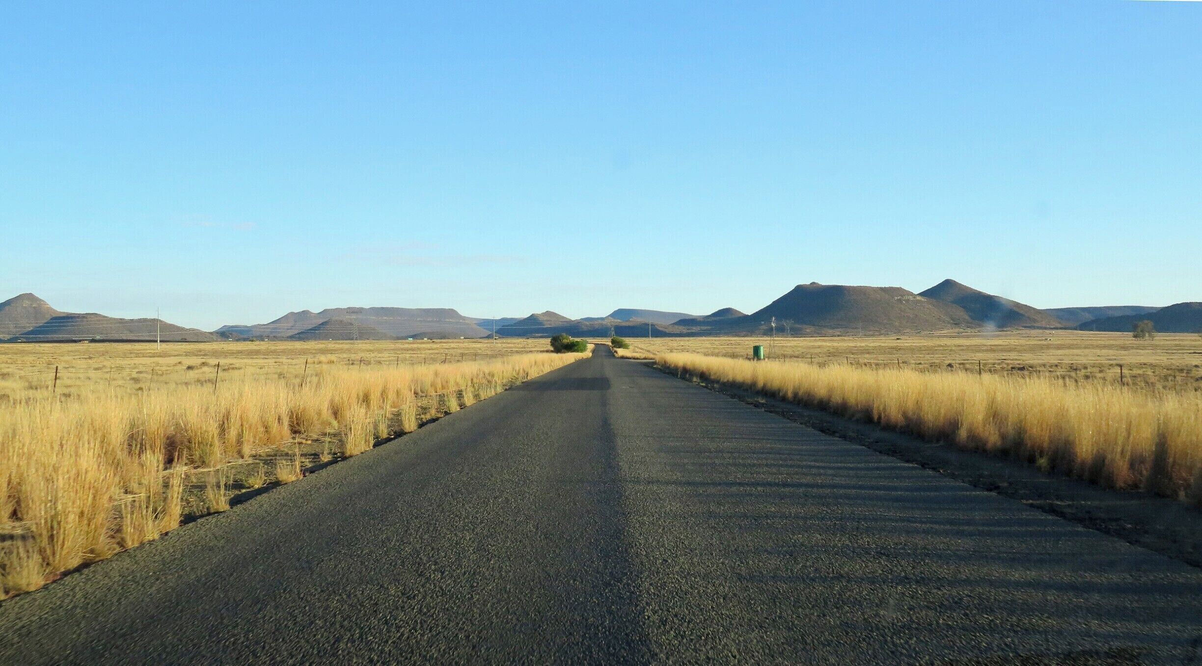 A straight road flanked by dry grass fields and distant mountains under a clear sky.