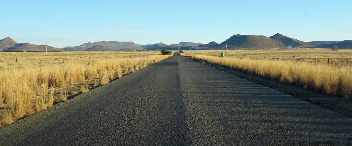 A straight road flanked by dry grass fields and distant mountains under a clear sky.
