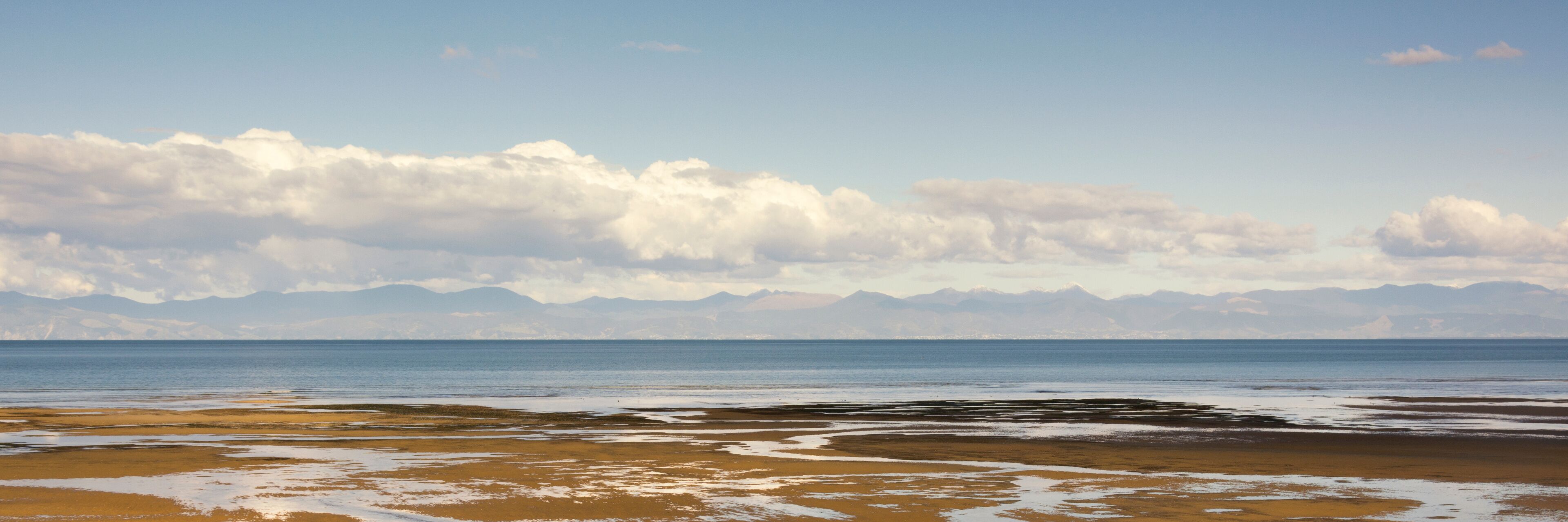 A beach with a calm sea, sandy shore, and distant mountains under a cloudy sky.