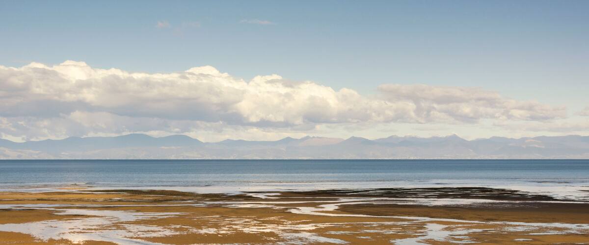 A beach with a calm sea, sandy shore, and distant mountains under a cloudy sky.