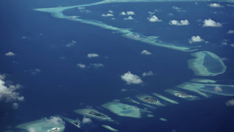 An aerial view of a chain of turquoise atolls with clear waters and scattered clouds.