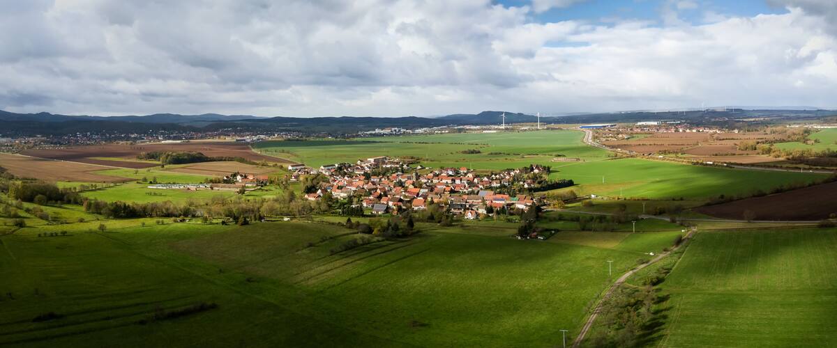 A rural landscape with rolling green fields, a small village with red-roofed houses, and distant wind turbines.