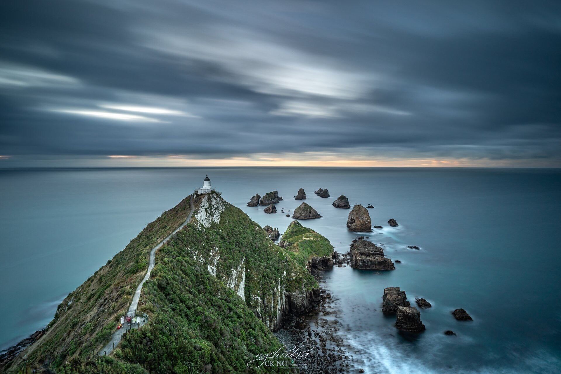 A coastal landscape with a lighthouse on a grassy hill, surrounded by rocky outcrops.