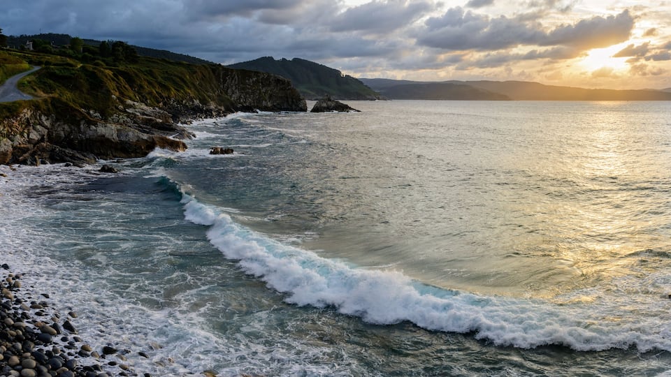 A coastal landscape with rocky shores, waves, and a winding road leading to a mountainous backdrop.