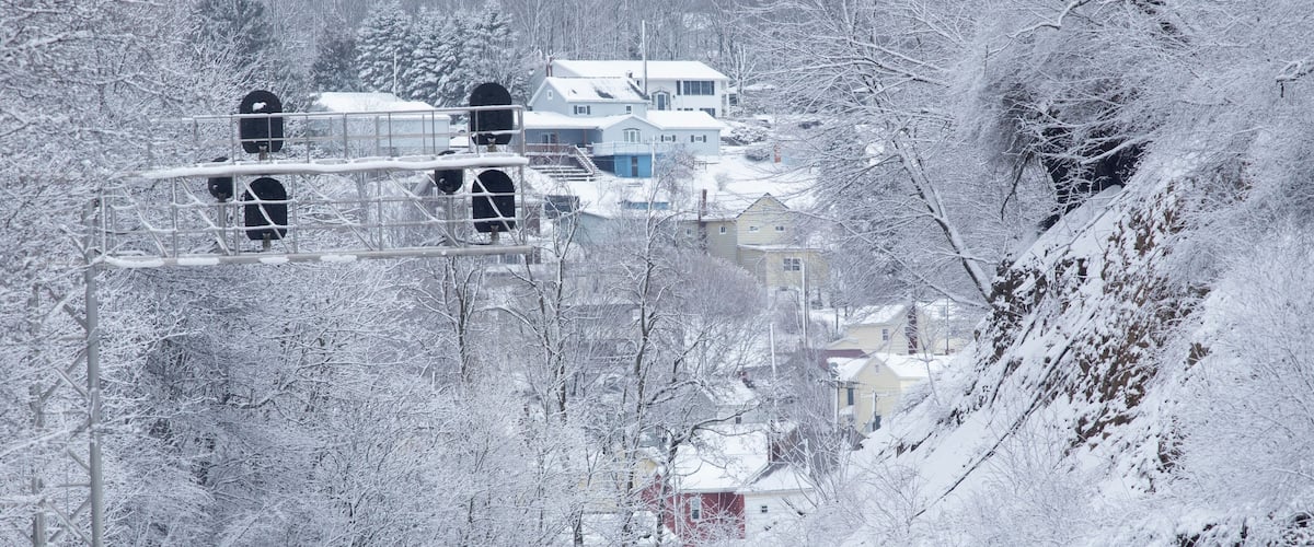 A snow-covered landscape with a railway track, a bridge, and houses in the background.