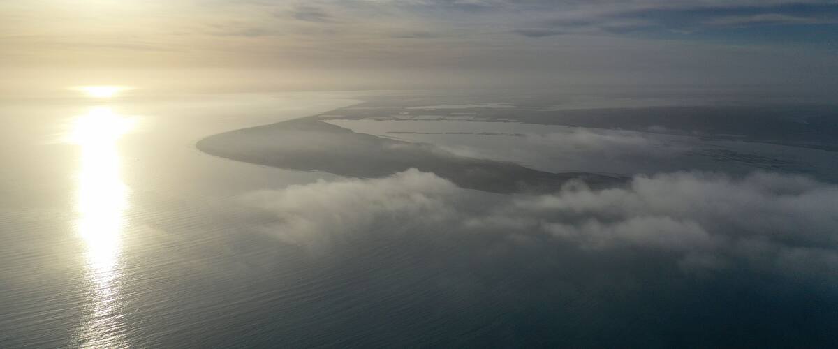 A coastal landscape with a sunlit sea, a cloud-covered island, and a clear sky.