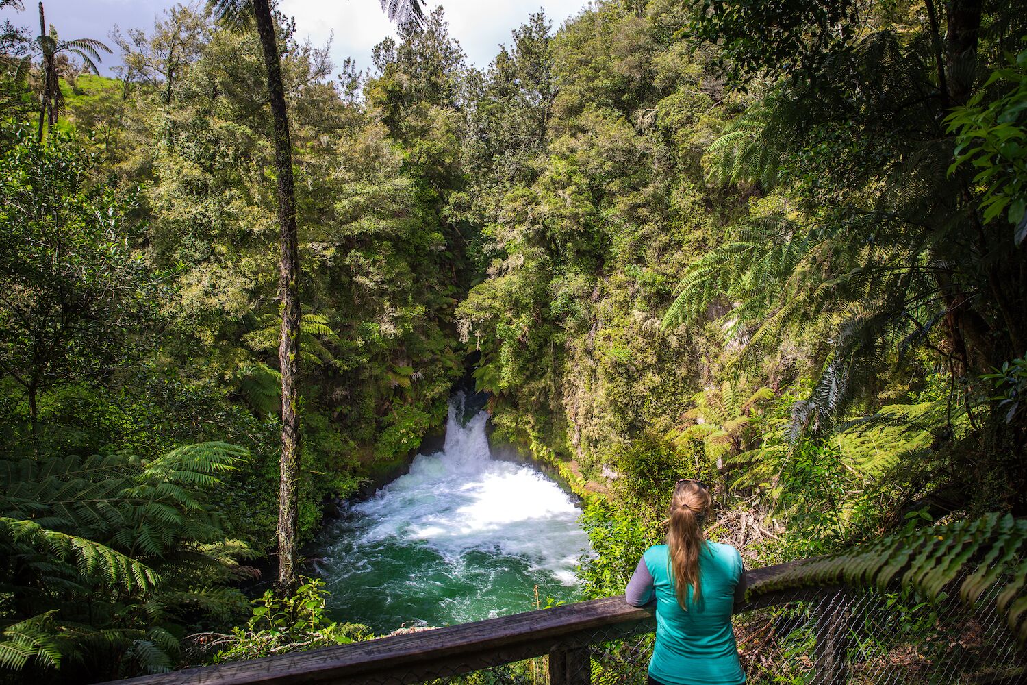 A woman standing on a wooden deck overlooking a waterfall in a lush green forest.