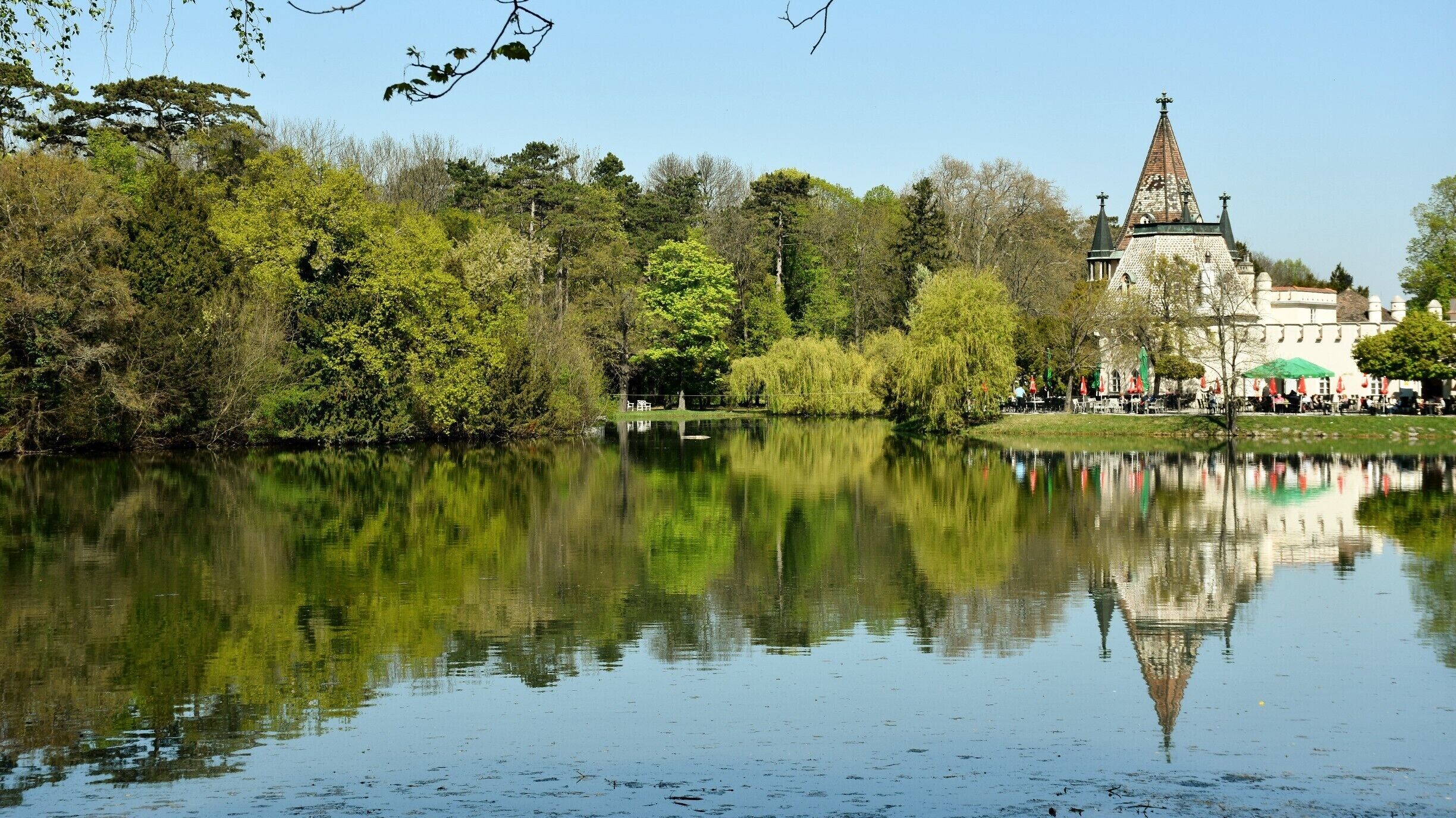 A calm lake reflecting trees and a historic building with a tall spire.