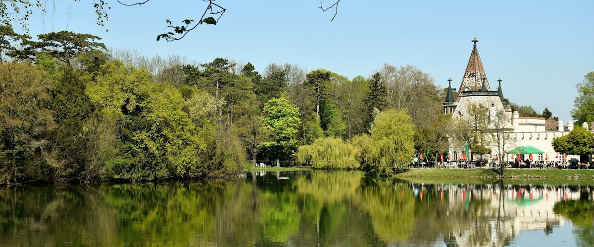 A calm lake reflecting trees and a historic building with a tall spire.
