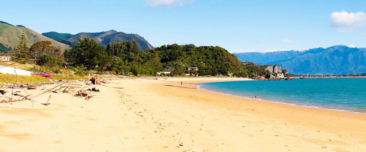 A sandy beach with clear turquoise water, surrounded by lush green hills and mountains in the background.