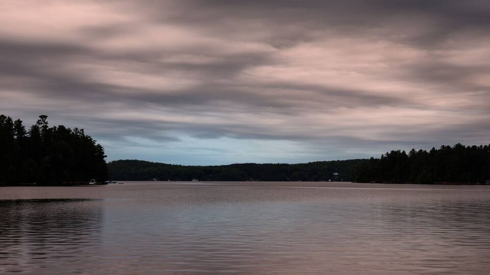A calm lake with a forested shoreline under a cloudy sky.
