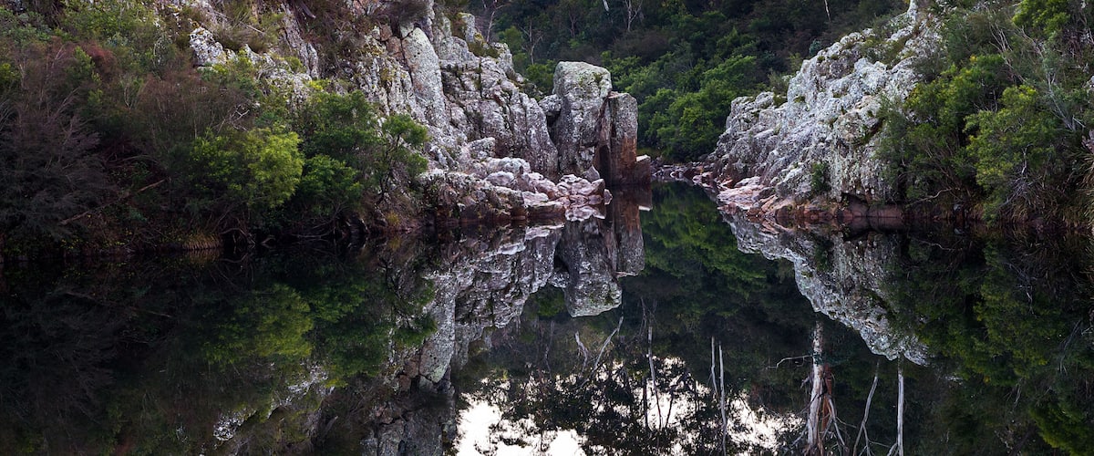 A rocky gorge with dense green foliage and a calm reflective water surface.