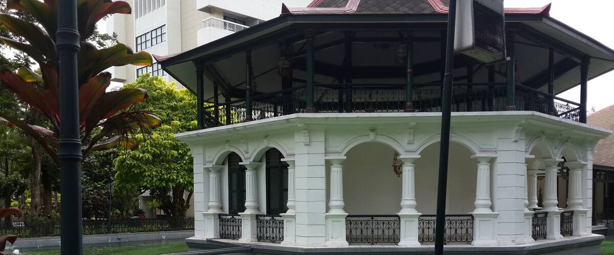 A white gazebo with arches and columns, surrounded by a greenish pond with a fountain.