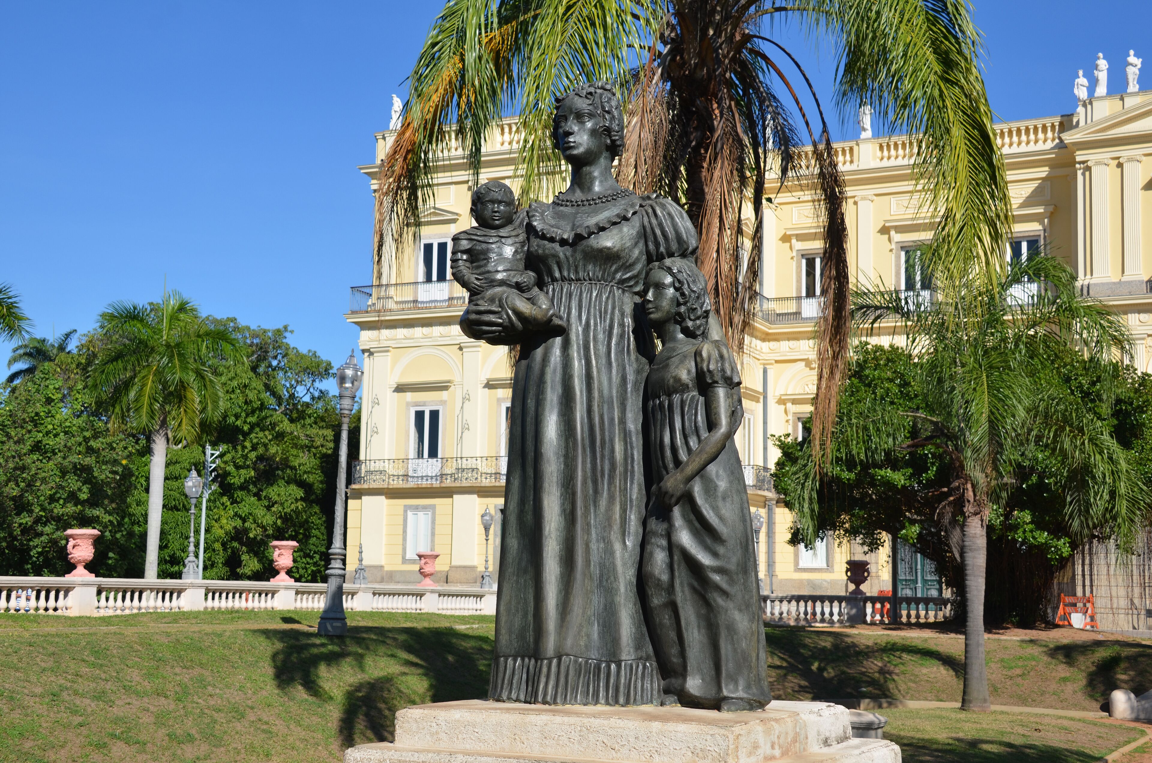A statue of a woman holding a child in a park with a large building in the background.