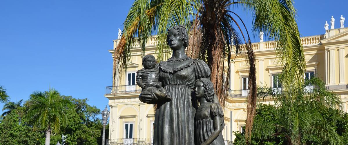A statue of a woman holding a child in a park with a large building in the background.