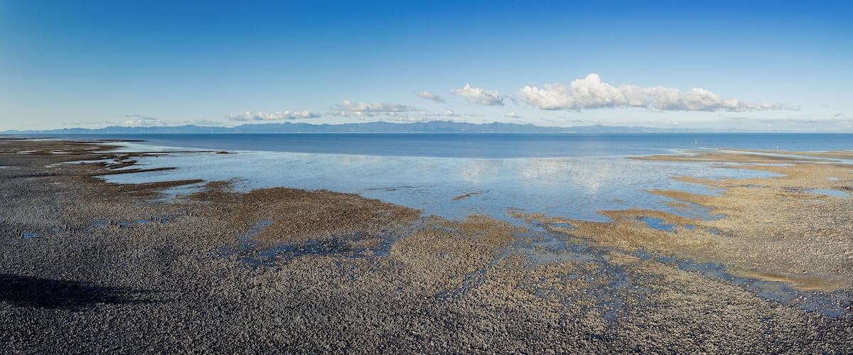 A pebbly beach with calm water and distant mountains under a clear sky.