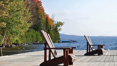Dos sillas Adirondack sobre un muelle de madera junto a un lago, con follaje otoñal y montañas al fondo.