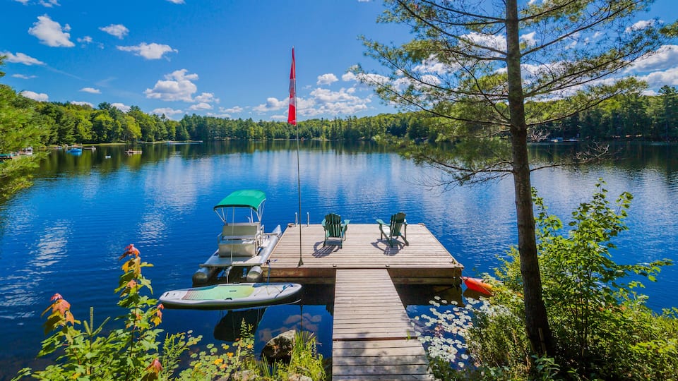 A calm lake with a wooden dock, green chairs, a small boat, and a red flag in the distance.