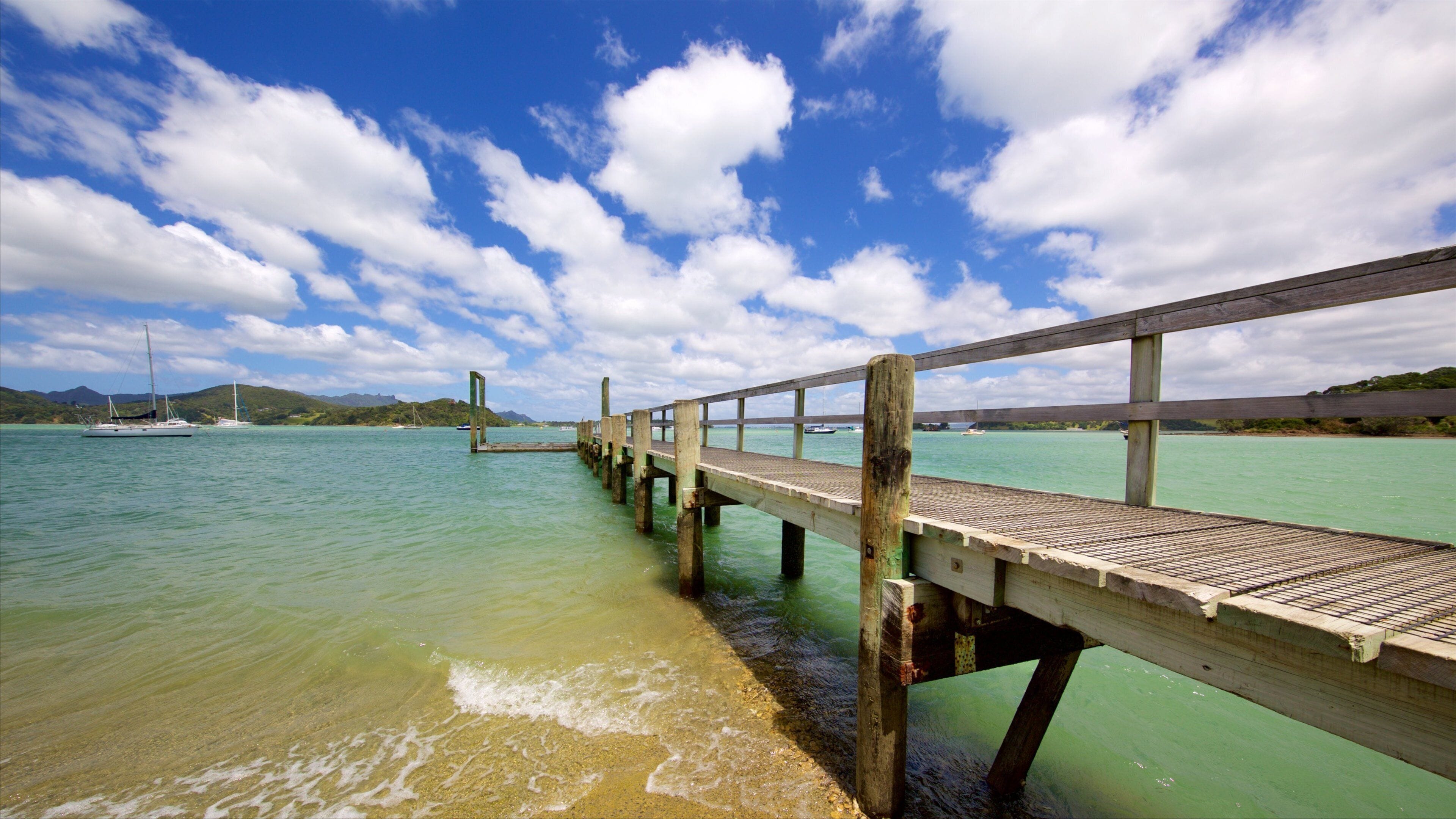 A wooden pier extending into turquoise waters with boats and a mountainous landscape in the background.
