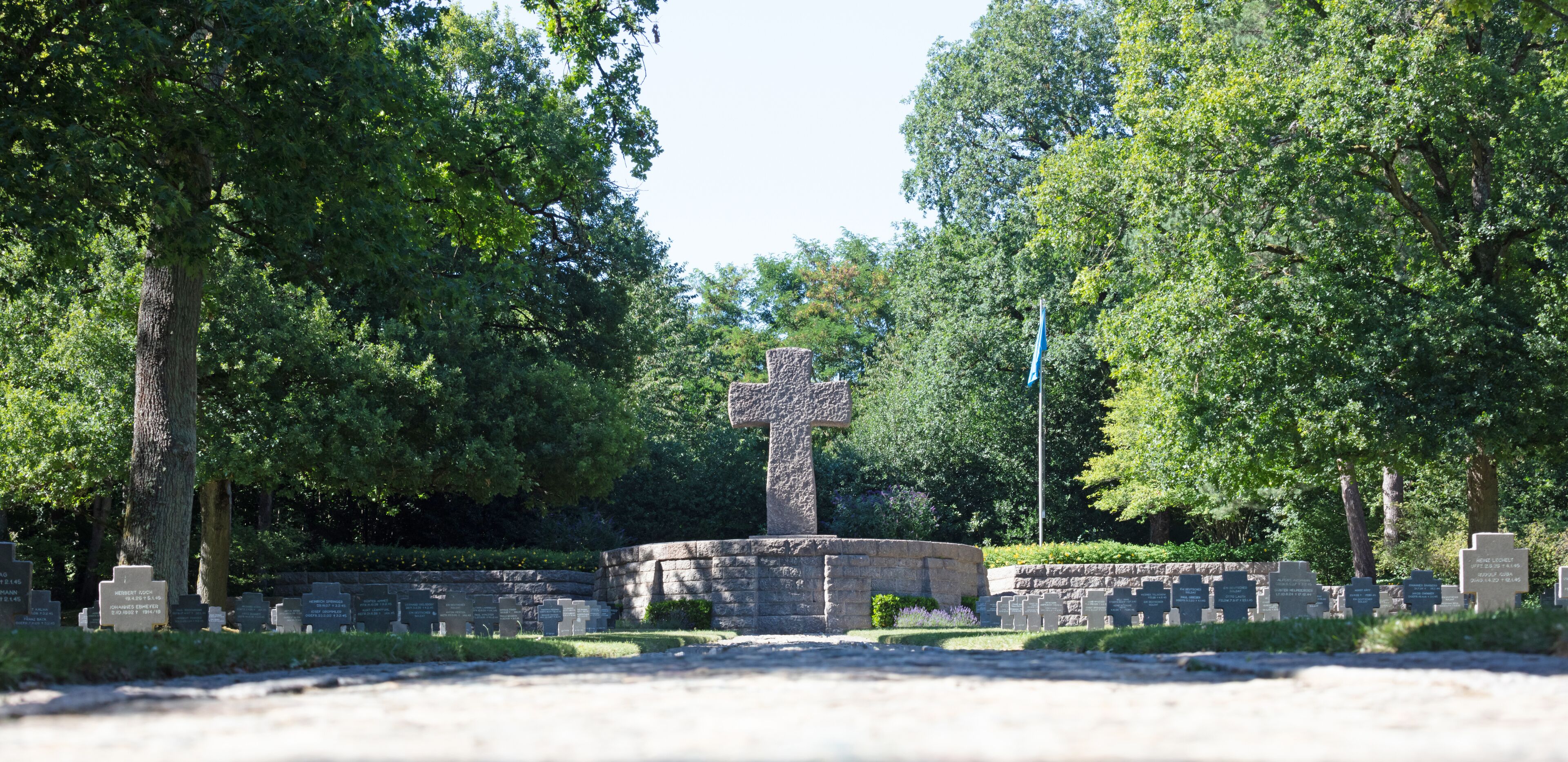 A cemetery with a prominent stone cross, surrounded by trees and numerous headstones.