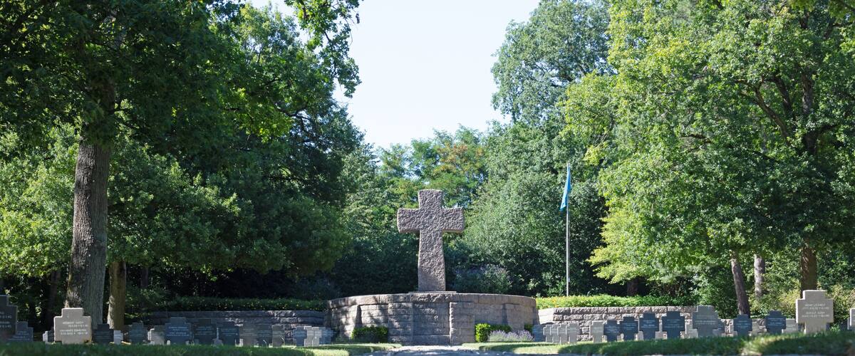 A cemetery with a prominent stone cross, surrounded by trees and numerous headstones.