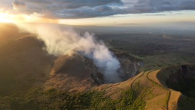 A volcanic crater with smoke rising, surrounded by a hilly landscape and a distant mountain range.