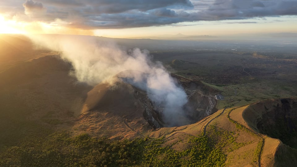 A volcanic crater with smoke rising, surrounded by a hilly landscape and a distant mountain range.