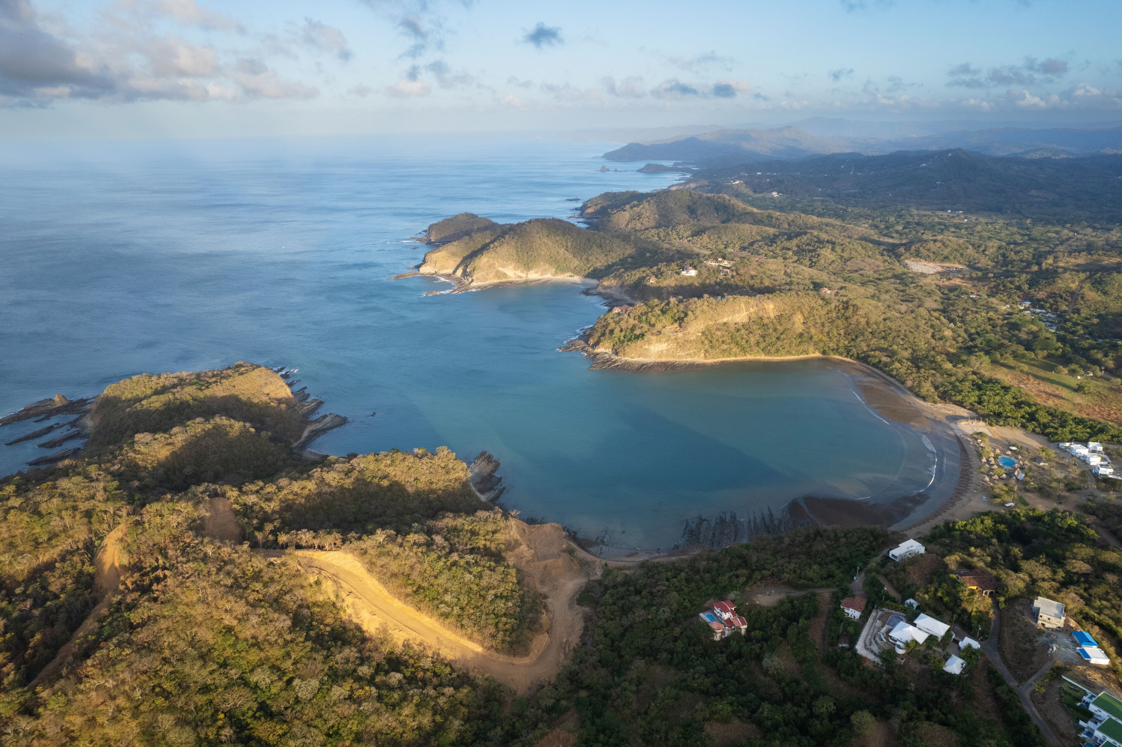 An aerial view of a coastal landscape with a large bay, forested hills, and scattered buildings.