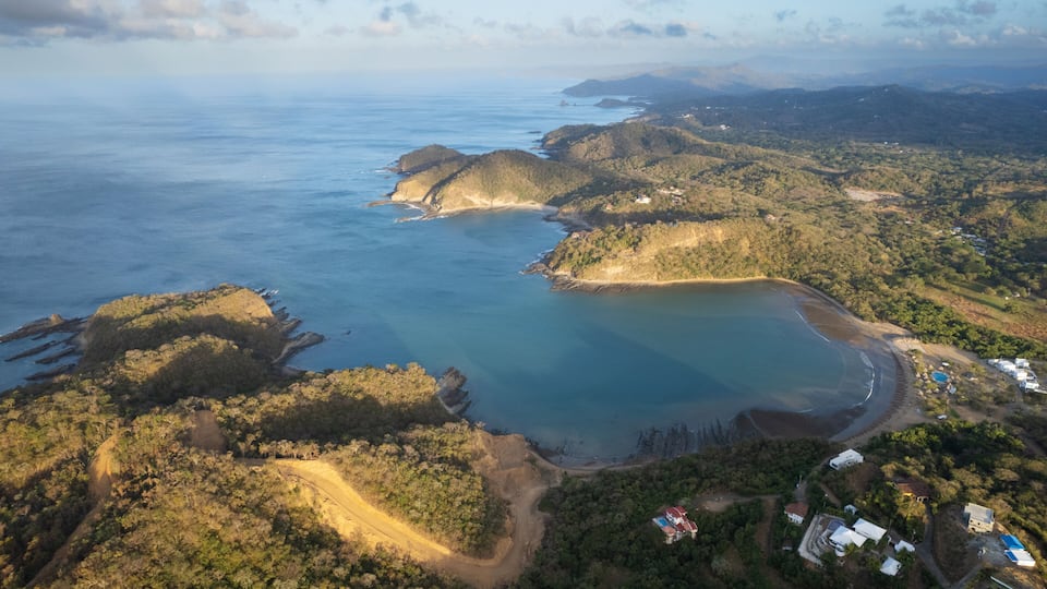 An aerial view of a coastal landscape with a large bay, forested hills, and scattered buildings.
