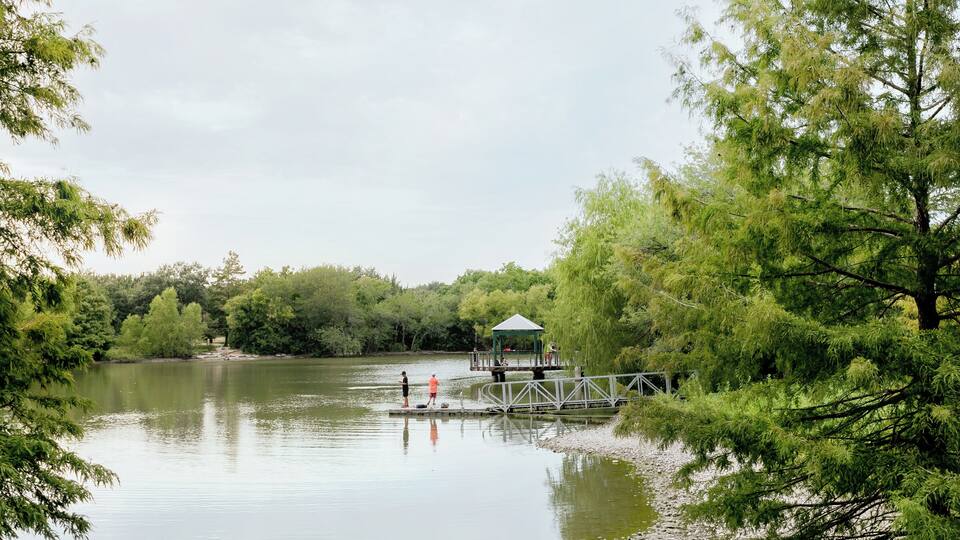 A calm lake with a wooden dock and gazebo, surrounded by dense green trees.