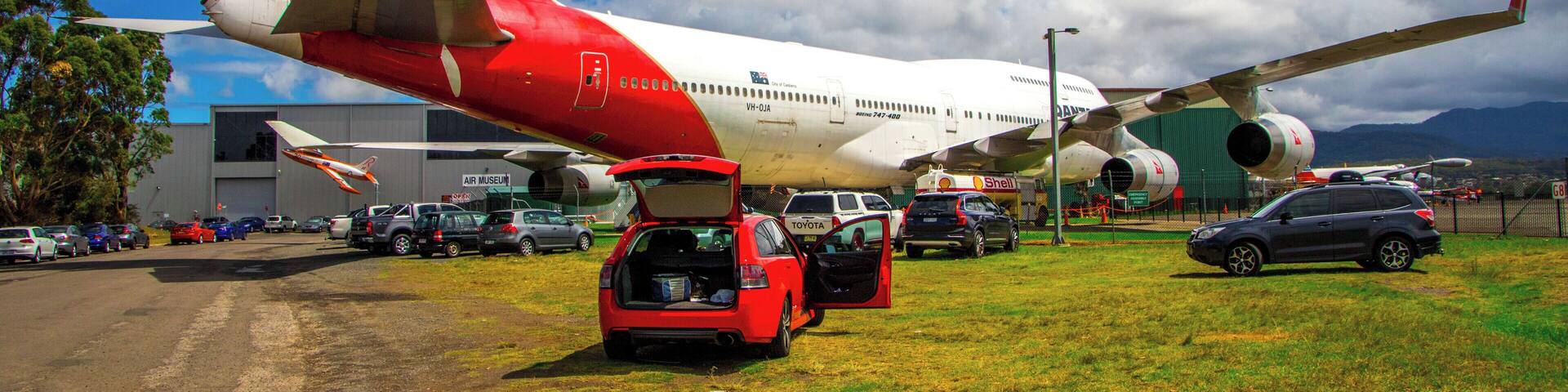 A large red and white airplane parked on a grassy field with cars and a building in the background.