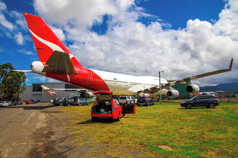 Un grand avion rouge et blanc est garé sur un champ d’herbe, avec des voitures et un bâtiment en arrière-plan.