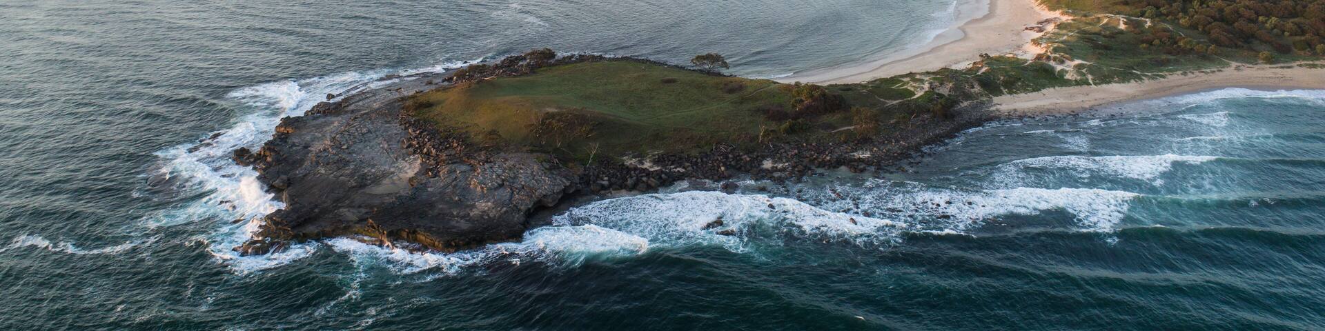 An aerial view of a coastal landscape with a sandy beach, rocky outcrops, and a forested area.