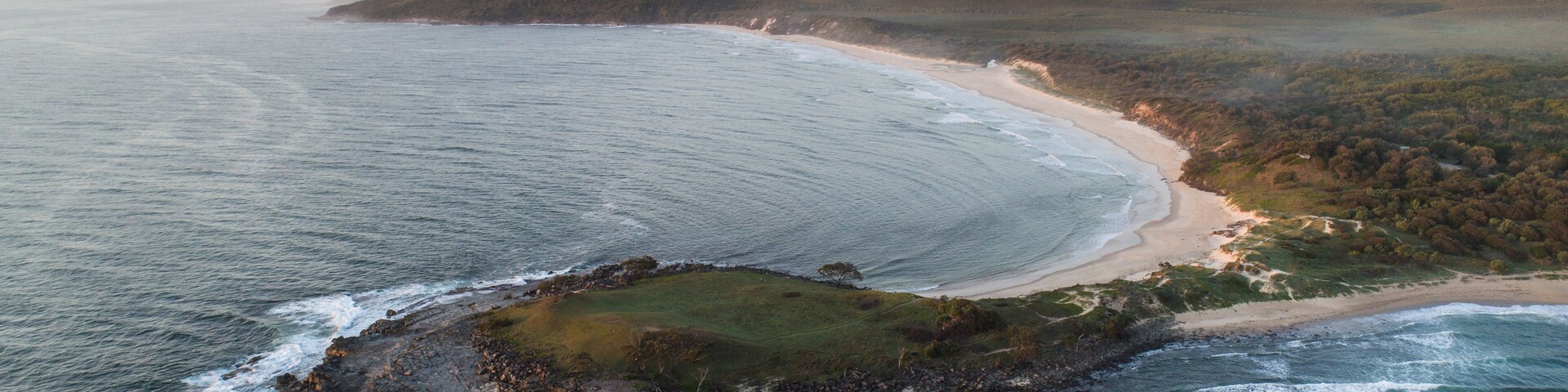 Vista aérea de un paisaje costero con una playa de arena, afloramientos rocosos y una zona boscosa.