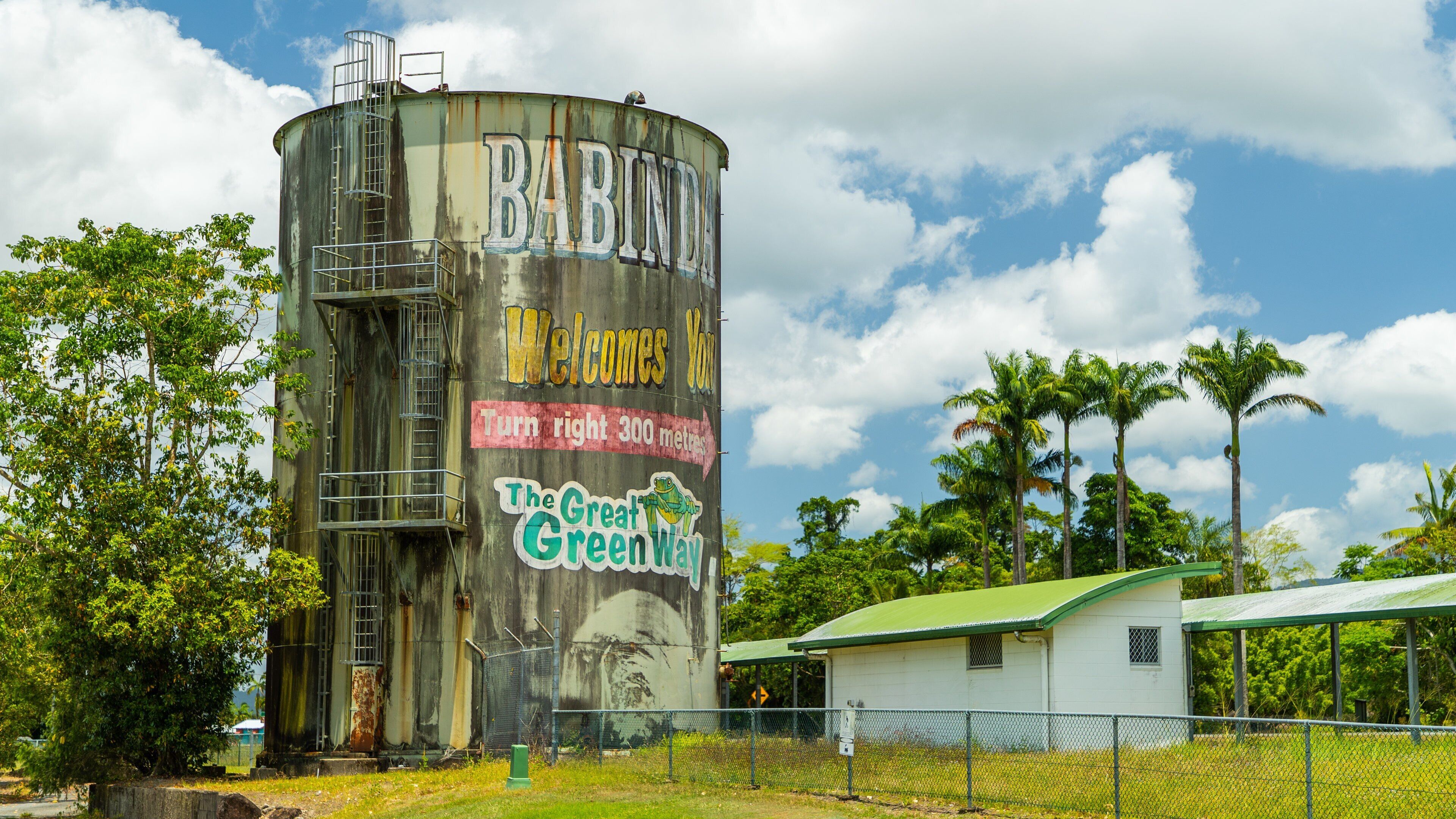 A large cylindrical water tank with graffiti, including 'Welcome You' and 'The Great Green Way', surrounded by trees and a small building.