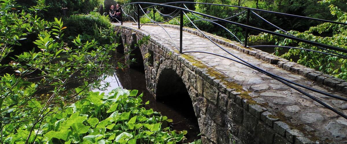 A stone bridge over a calm water body, surrounded by lush greenery and trees.