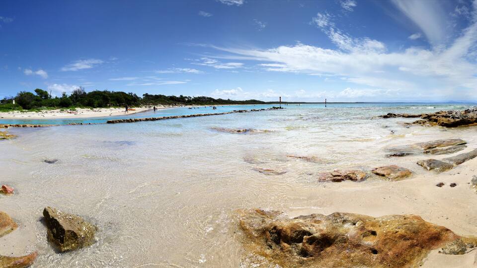 A beach with clear water, rocks, and a distant shoreline with trees and buildings.
