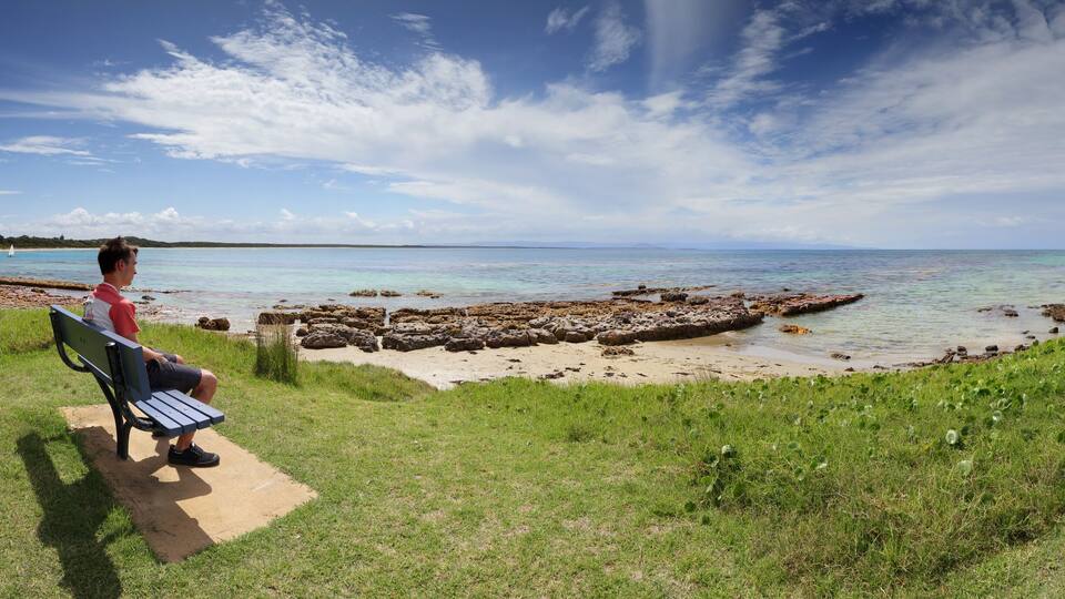 A man sitting on a bench overlooking a beach with clear water and rocky outcrops.