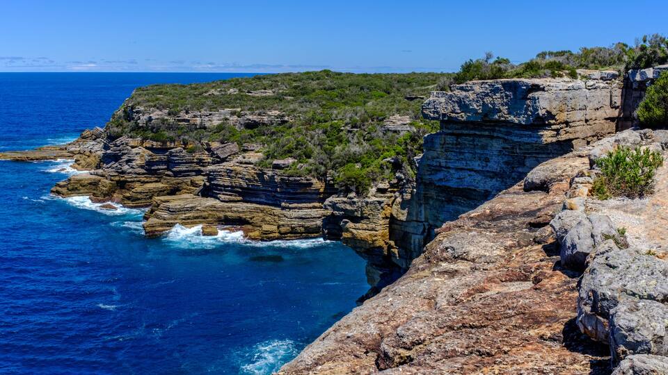 A coastal cliff with layered rock formations and a clear blue sea.