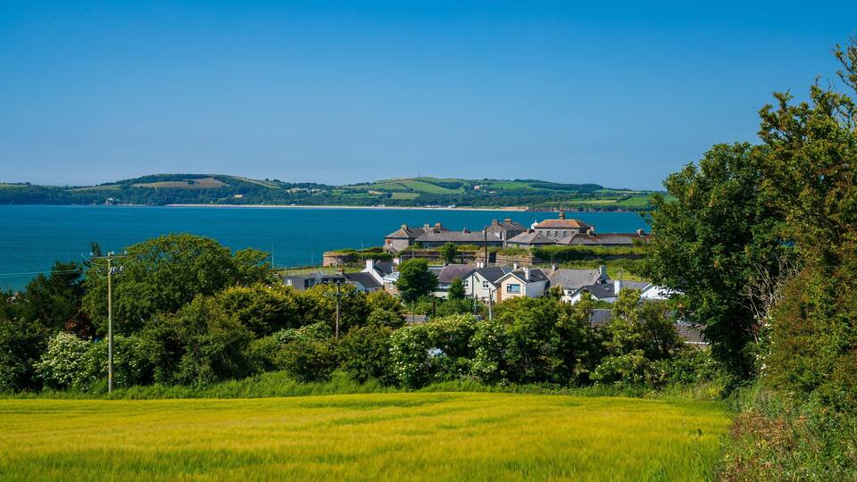 A coastal village with houses, a field, and a view of the sea and hills.