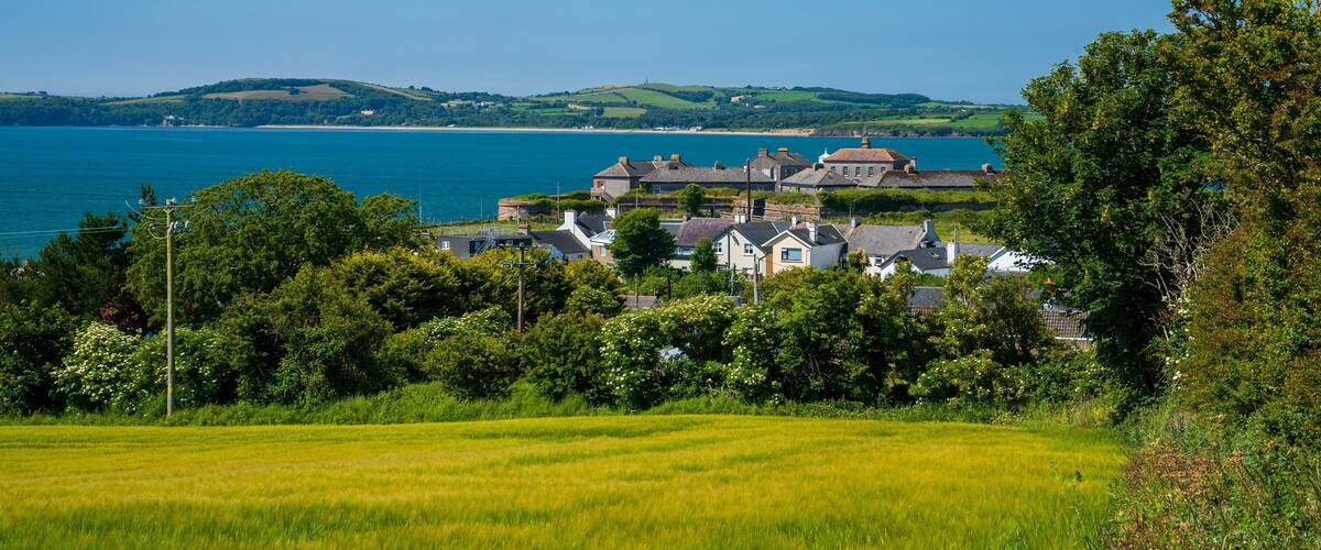 A coastal village with houses, a field, and a view of the sea and hills.