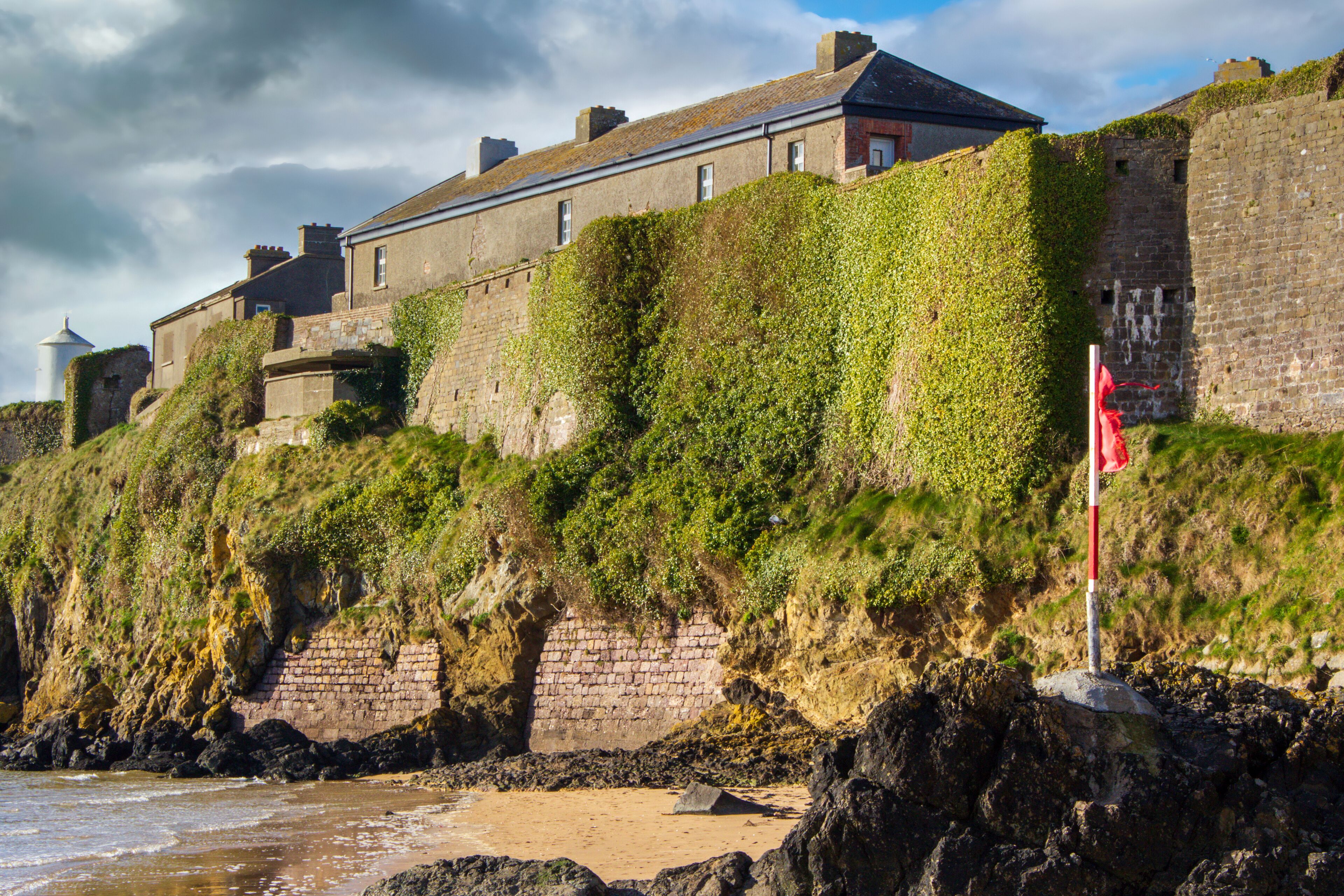 A coastal castle with ivy-covered walls, a flagpole, and a lighthouse in the background.