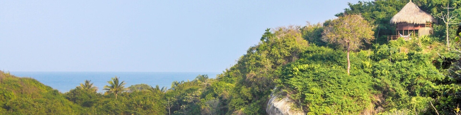 Uma praia com ondas, guarda-sóis e um edifício com telhado de palha numa falésia.