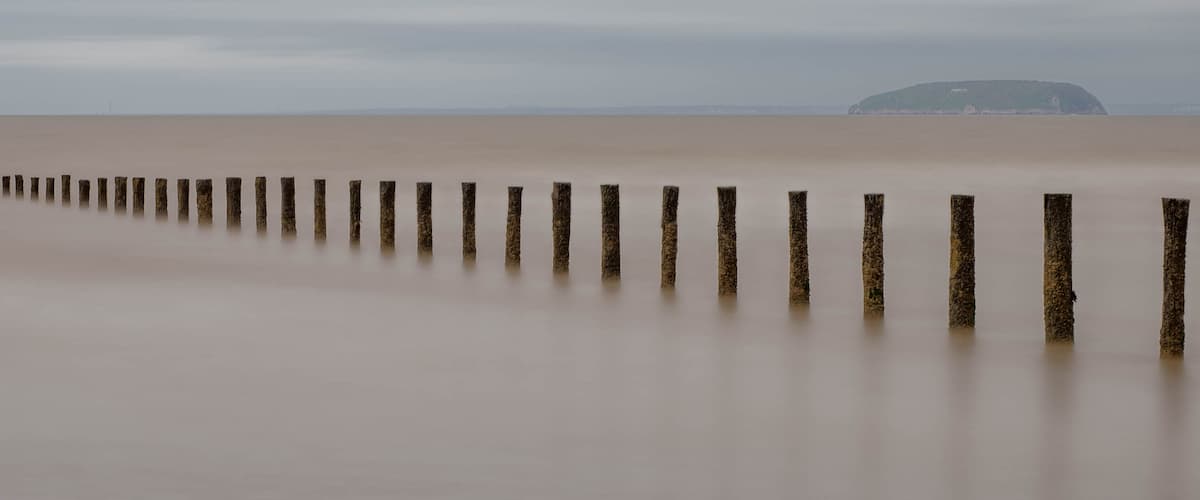 A long, straight wooden breakwater extending into calm waters, with a distant hill and overcast sky.