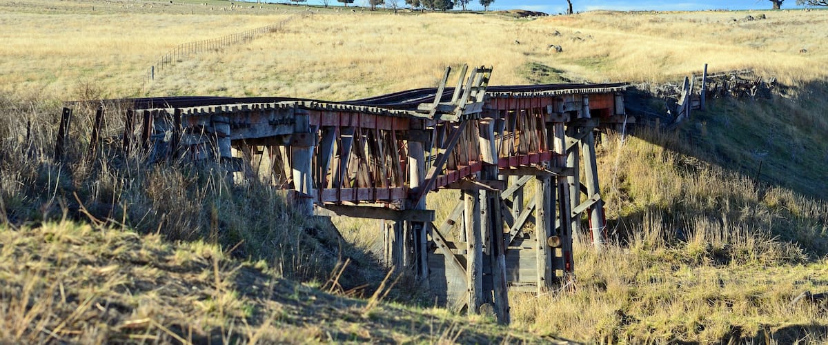 A rusted railway bridge in a grassy field with trees in the background.