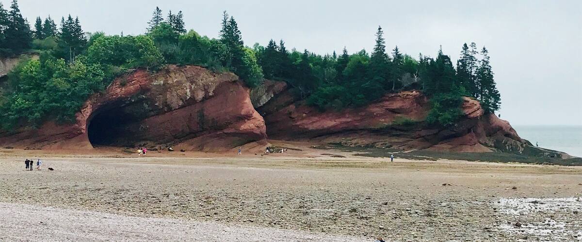 A rocky cliff with a cave entrance, a beach, and people walking.