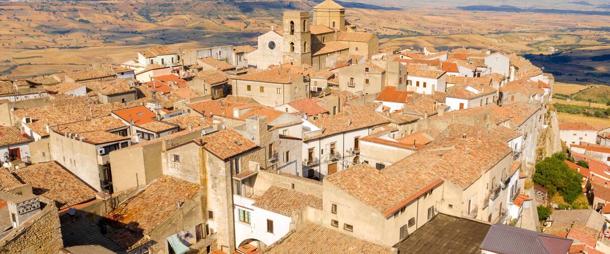 A historic town with terracotta-roofed buildings, a prominent church with a bell tower, and rolling hills in the background.