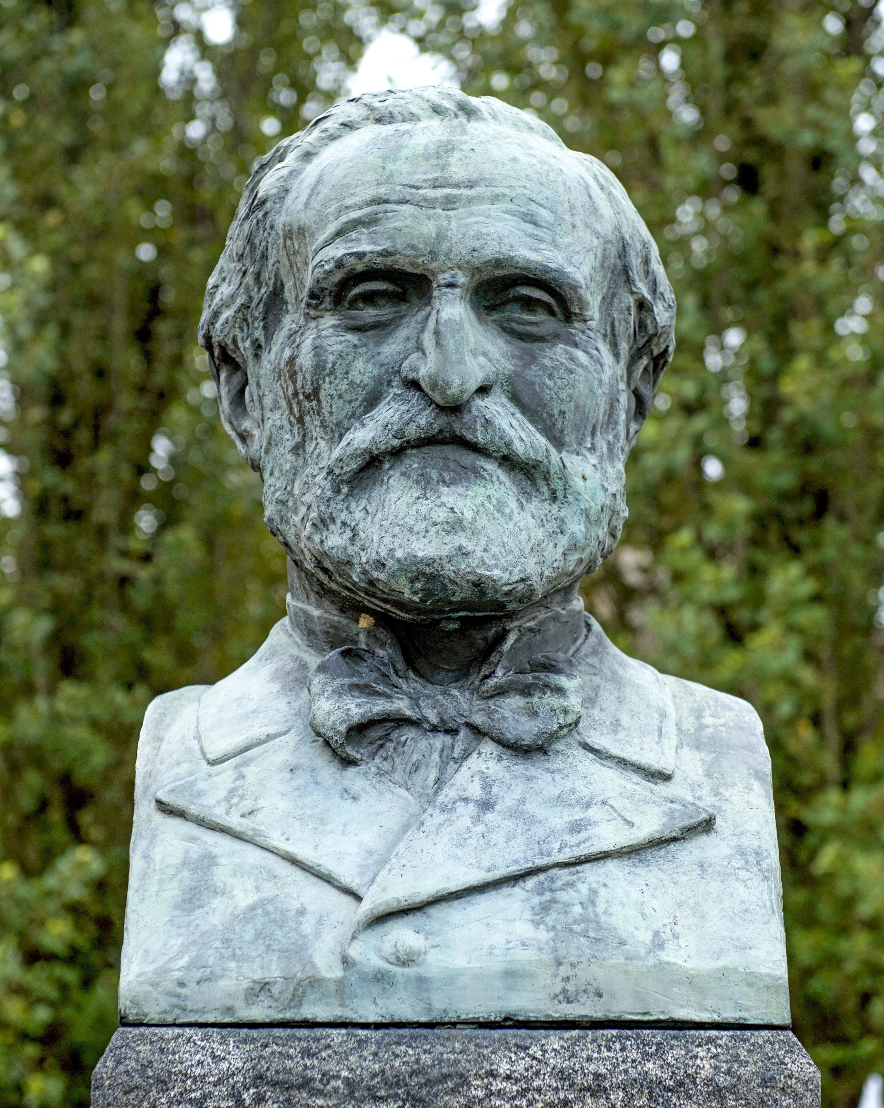 A statue of a man with a bow tie and a suit, set against a backdrop of trees.