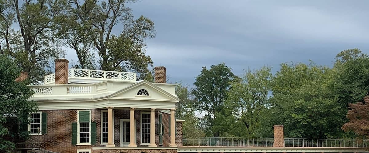 A historic brick building with a white porch and columns, surrounded by a grassy field and trees.